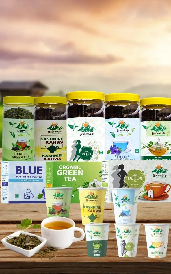 A display of Brahmkalp tea products, including various jars, boxes, and cups featuring herbal green tea, Kashmiri Kahwa, detox tea, butterfly pea tea ("Blue"), and organic green tea, arranged on a wooden table with a scenic sunset background